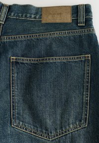 Vue arrière d'un jean en denim bleu foncé montrant une seule poche arrière avec des coutures contrastantes et une étiquette en cuir marron indiquant "DENIM SUPPLY".