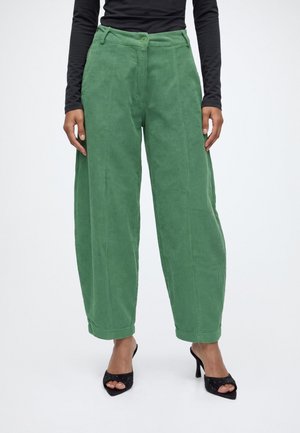 Modèle portant un pantalon en velours côtelé vert à jambes larges, avec une taille boutonnée et des sandales noires à talons.