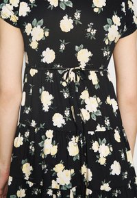 Svart blommig klänning i mjukt tyg, med gula rosor, gröna blad och ett knytband i midjan samt volangkant i lager.