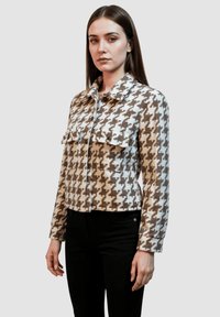 Chaqueta de cuadro houndstooth en marrón y blanco con cuello tipo solapa, dos bolsillos en el pecho y un diseño entallado. Combinada con pantalones negros.