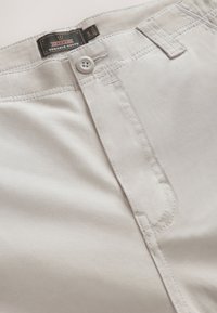 Pantalon beige clair avec fermeture par bouton, passants de ceinture et étiquette "Next Durable Goods" à l'intérieur de la ceinture.