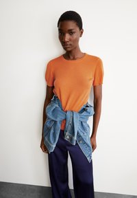 Oranje gebreide top met korte mouwen, gecombineerd met een spijkerjack dat om de taille is geknoopt. Donkere wijde broek maakt de outfit compleet.