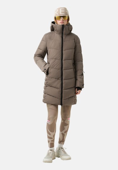 Gepolsterter Mantel in Taupe mit hohem Kragen, Reißverschluss vorne und Seitentaschen. Kombiniert mit beigen Camouflage-Leggings und klobigen Sneakers.