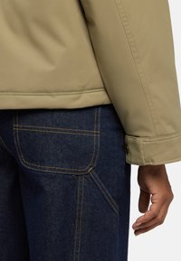 Giacca beige con texture liscia e design strutturato, abbinata a jeans in denim blu scuro con cuciture arancioni e tasche laterali.