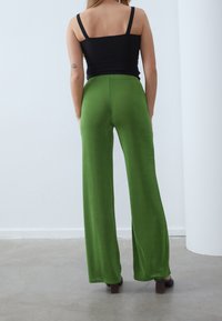 Kvinna som står inomhus klädd i gröna vida byxor, svart ärmlös topp och svarta klackskor, sedd bakifrån.