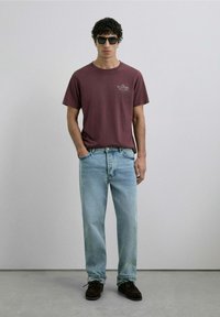 Camiseta de cuello redondo color burdeos con logo, jeans rectos de color azul claro, zapatos de ante marrón y gafas de sol negras. Fondo liso, vista frontal.