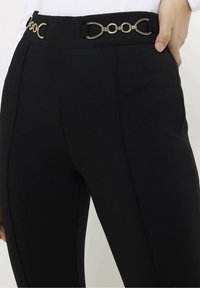 Zwarte broek met een gladde textuur, voorzien van een decoratieve gouden kettingriem bij de taille. Slim-fit ontwerp met op maat gemaakte details.