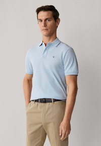 Homme debout, les mains dans les poches, portant un polo bleu clair et un pantalon beige, devant un fond clair uni.