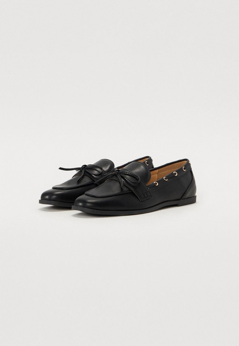 Scarpe in pelle nera con punta tonda, dettaglio a fiocco decorativo, interno contrastante color crema e occhielli metallici per i lacci. Design con suola piatta.