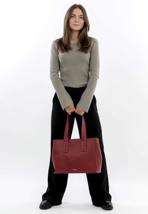 Sac fourre-tout en simili cuir rouge avec finition texturée, forme rectangulaire, double poignées et détail de logo métallique. Porté avec un pantalon noir.