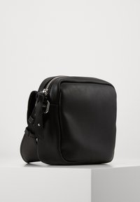 Kompaktní černá kožená crossbody taška se stříbrným zipem a nastavitelným popruhem, ležící na bílém podstavci na jednobarevném pozadí.
