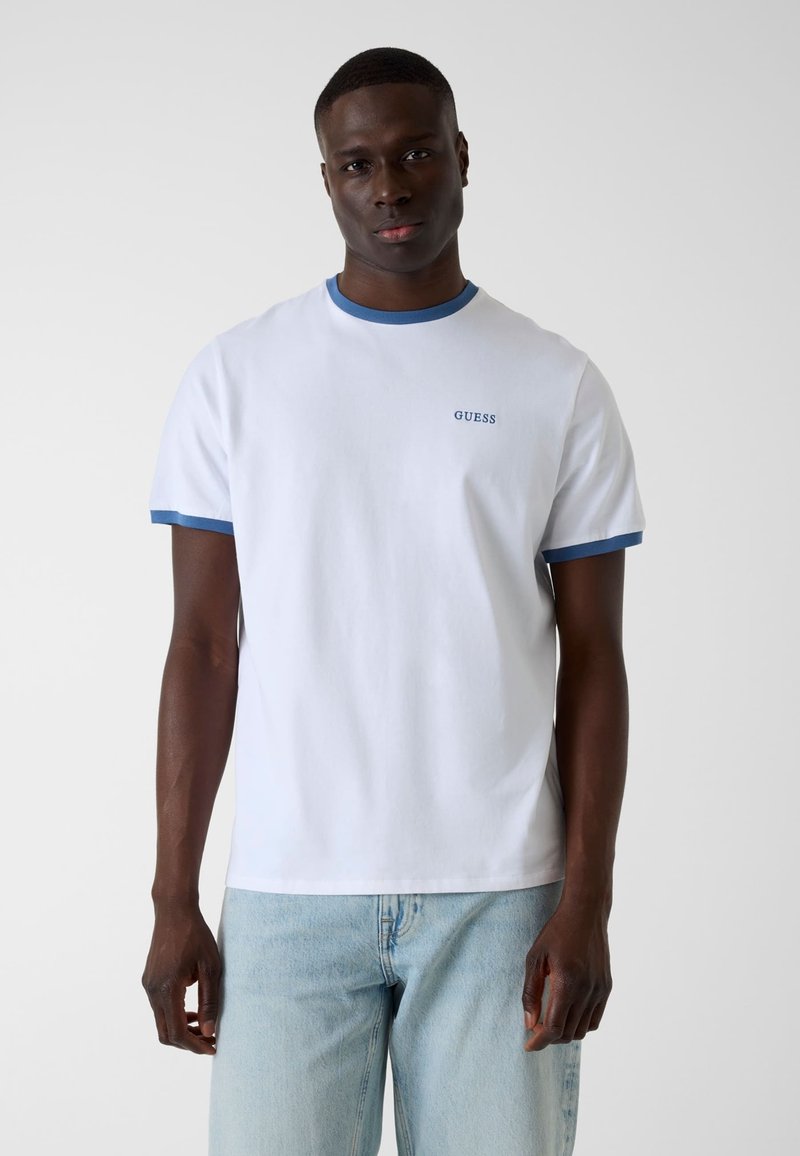 Homme portant un T-shirt blanc avec bordure bleue et logo "GUESS", associé à un jean bleu clair, debout devant un fond uni.