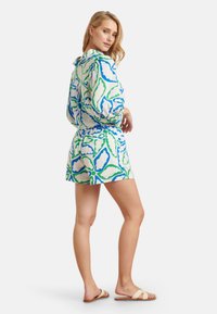 Korte mouw romper met patroon in groen en blauw op een witte achtergrond, met een getailleerde taille en een lusdetail aan de achterkant. Platte sandalen gedragen.