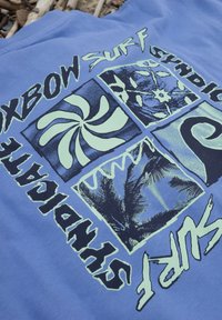 Tissu bleu avec un imprimé de quatre carrés montrant des formes abstraites, des palmiers et des vagues, avec les mots "Oxbow Surf Syndicate".