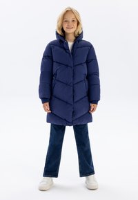 Abrigo acolchado azul marino con un patrón de rombos, capucha y puños acanalados, combinado con jeans de campana y zapatillas blancas.