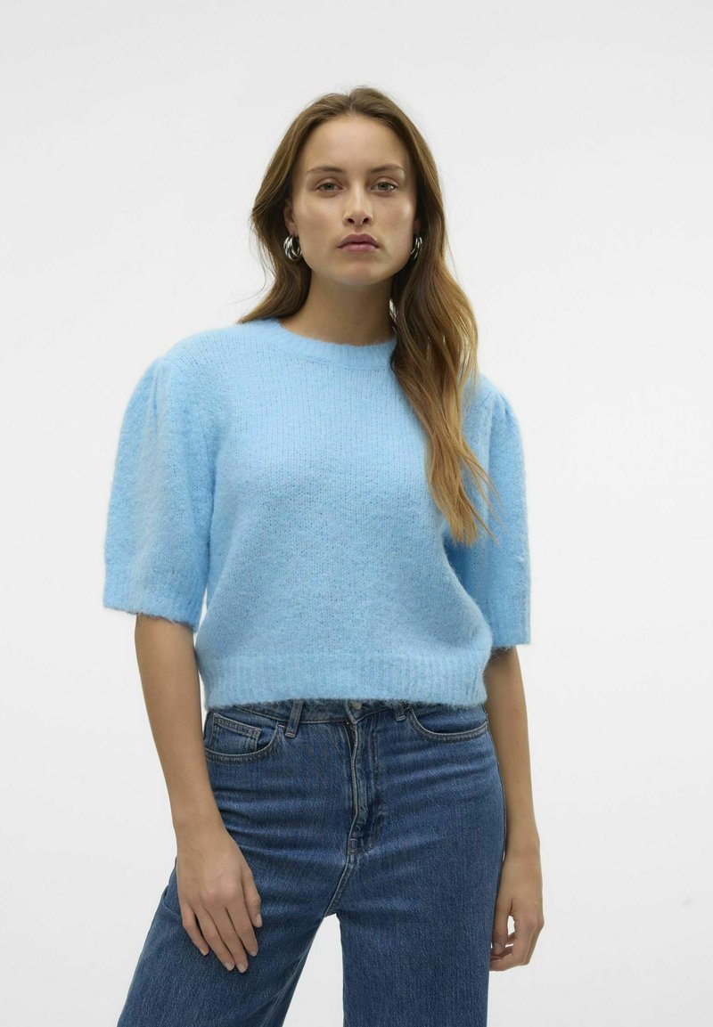 Gaiši zils, īsām piedurknēm adīts džemperis no mīksta, pūkaina materiāla ar apaļu kakla izgriezumu un brīvu piegriezumu, apvienots ar ziliem džinsiem.