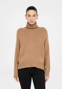 Donna che indossa un maglione a collo alto color cammello con polsini e orlo a costine, pantaloni neri e orecchini a forma di cuore dorati, in piedi di fronte a uno sfondo bianco.