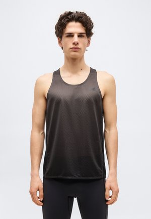 Mladý muž s kučeravými vlasmi, oblečený v čiernom atletickom tielku bez rukávov a čiernych legínach, stojaci pred jednoduchým svetlým pozadím.