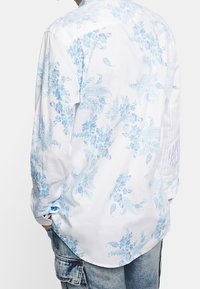 Camicia a maniche lunghe in cotone bianco con motivi paisley e floreali azzurri. Presenta un colletto button-down e maniche arrotolate con dettagli a righe.