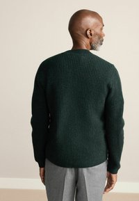 Dunkelgrüner gerippter Strickpullover mit rundem Ausschnitt, von hinten gezeigt, mit langen Ärmeln und einer subtilen Textur. Graue Hose vervollständigt das Outfit.