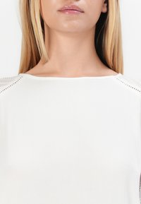 Weißes Oberteil mit rundem Ausschnitt und strukturierten Spitzelementen an den Schultern. Weicher Stoff mit glatter Oberfläche. Ärmel in mittlerer Länge.