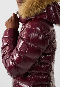 Burgunderfarbene Pufferjacke mit glänzender Oberfläche, mit Fell gefütterter Kapuze, gestepptem Design und Logopatch am Ärmel. Die Textur wirkt weich und warm.