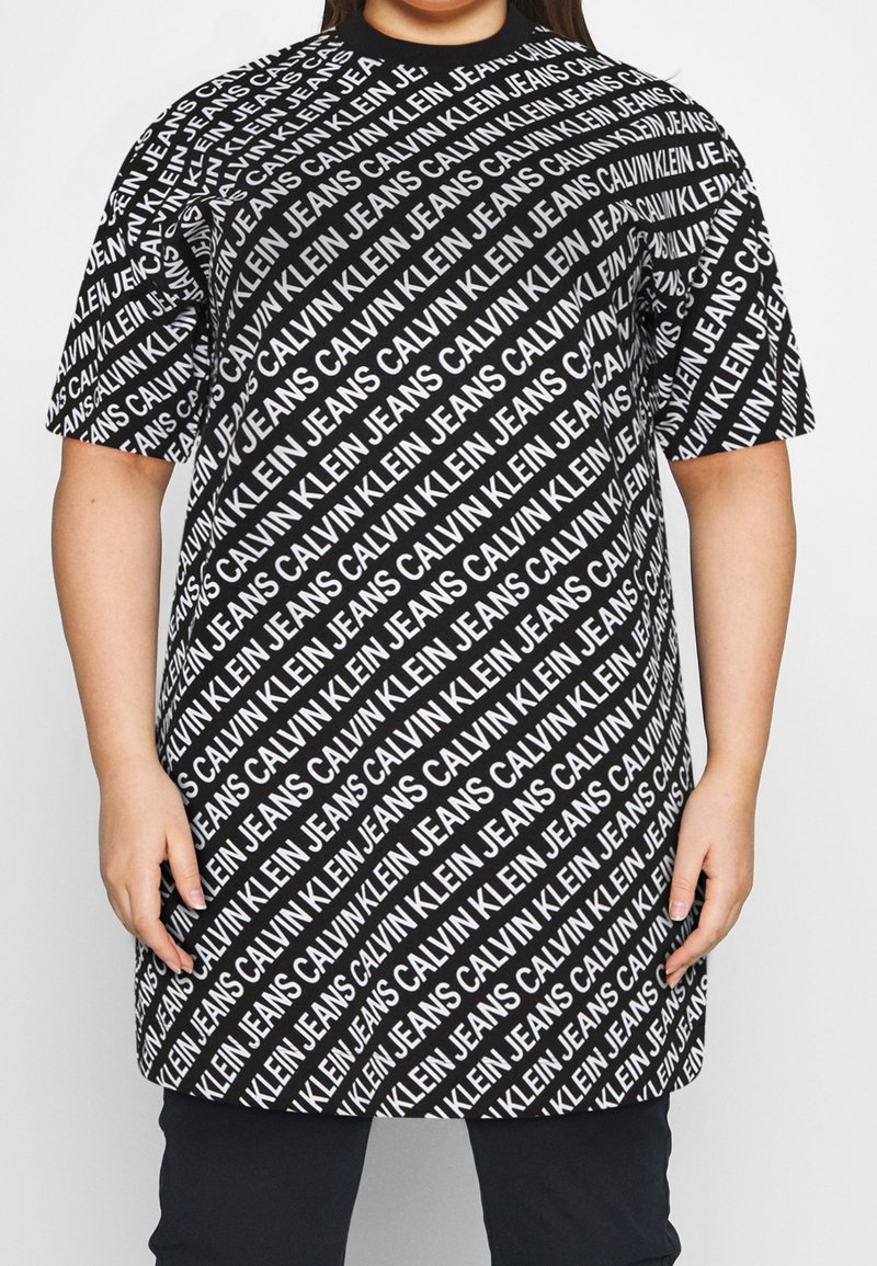 Svart oversize T-shirt med ett upprepat vitt textmönster av "CALVIN KLEIN JEANS." Korta ärmar och rund halsringning.