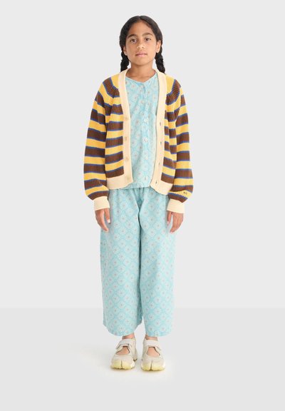 Fille aux cheveux tressés portant un pyjama bleu à motif, un cardigan rayé jaune et marron, et des baskets blanches, debout devant un fond uni.