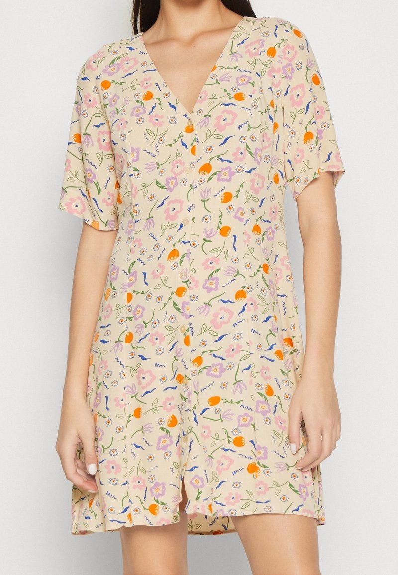 Femme portant une robe courte beige à col en V et manches courtes, recouverte d'un imprimé floral coloré avec des accents roses, orange et bleus.