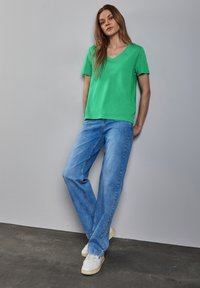 Mladá žena se opírá o šedou zeď, má na sobě zelené tričko s výstřihem do V, světle modré džíny a bílé tenisky.