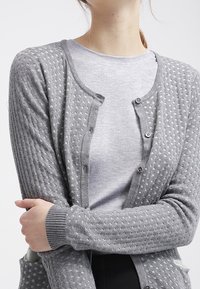 Grauer Strickcardigan mit strukturiertem Muster, Knopfverschluss und gerippten Bündchen. Über einem hellgrauen Hemd getragen für einen geschichteten Look.
