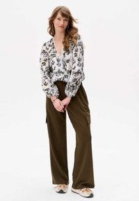 Weißbluse mit floralem Muster in Grün und Lila; lockere, weit geschnittene Ärmel; kombiniert mit olivgrünen, weit geschnittenen Hosen; weicher Stoff; lässige Sneakers.