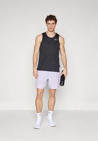 Mladý muž v čiernom bezrukávovom tričku Nike, svetlofialových šortkách, bielych ponožkách Nike a topánkach, drží čiernu fľašu na vodu a stojí proti jednému pozadiu.