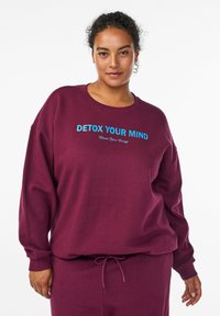 Bordová mikina z mäkkej látky s voľným strihom. Má modrý nápis "DETOX YOUR MIND" a "Create Your Energy." Sťahovacia šnúrka v oblasti pásu.