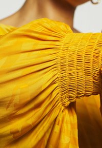 Yellow garment detail featuring smocking on the sleeve, with a soft, textured fabric and subtle pattern, highlighting a voluminous design.