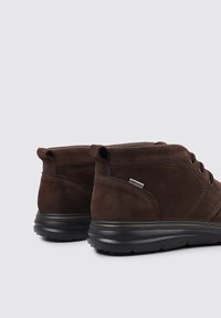 Un par de botas de cuero marrón con cordones hasta el tobillo y suelas de goma negras sobre un fondo gris claro, mostradas desde la parte posterior.