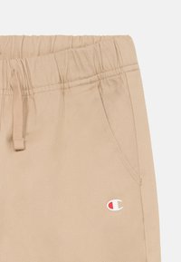 Pantalons beiges décontractés avec taille élastique, cordon de serrage, poches avant et petit logo Champion près d'une poche.
