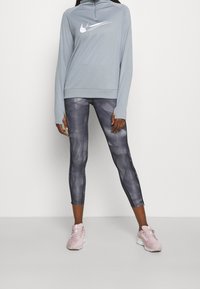 Felpa azzurra con zip e logo bianco, abbinata a leggings grigi scuri con motivo. Indossata con scarpe da corsa rosa chiaro.