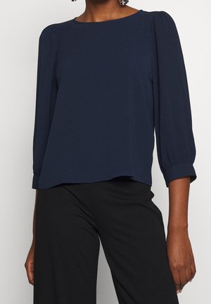 Femme portant un chemisier bleu marine à manches bouffantes trois-quarts et un pantalon noir à jambes larges sur un fond uni.