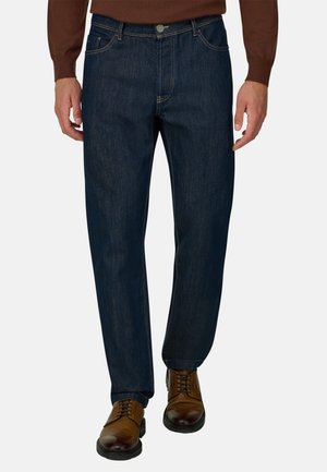 Homme portant un jean bleu foncé coupe droite, des chaussures habillées marron et une chemise marron à manches longues, debout devant un fond uni.