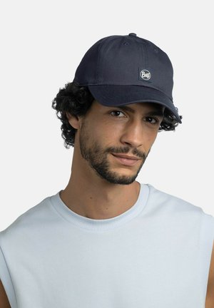 Mann mit dunklem lockigem Haar und Bart, der eine schwarze Buff-Mütze und ein hellblaues ärmelloses Oberteil trägt, schaut mit leichtem Lächeln nach vorne.