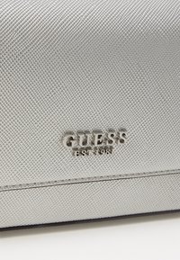 Srebrna torebka o teksturze z geometrycznym wzorem oraz błyszczącą logo "GUESS EST. 1981" z przodu.