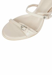 Sandalo con tacco alto beige in pelle liscia, con sottili cinturini alla caviglia e un piccolo dettaglio in metallo sulla cinghia.