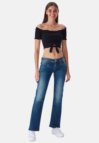 Top corto negro sin mangas con detalle de nudo en la parte delantera, combinado con jeans azules acampanados. La modelo lleva zapatillas blancas. Fondo simple.