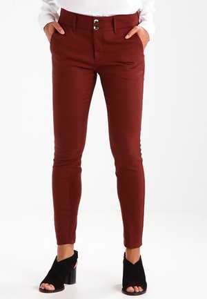 Frau trägt schmal geschnittene, rostfarbene Hose mit aufgesetzten Fronttaschen und Doppelknopfverschluss, kombiniert mit schwarzen, offen zehigen Blockabsatz-Schuhen.