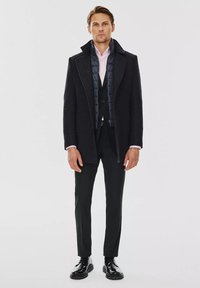 Męski płaszcz z ciemnego materiału o strukturalnym wzorze. Posiada pikowaną podszewkę, szerokie klapy, dopasowany krój oraz czarne spodnie formalne i buty.