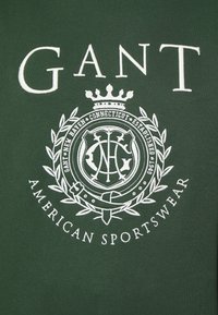 Sudadera verde con texto bordado en blanco "GANT" y un emblema circular con acentos de laurel, que detalla "AMERICAN SPORTSWEAR".