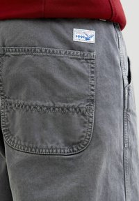 Grå denimshorts med en stor baglomme og et blått og hvitt mærke med en ørnegrafik og teksten "1990".