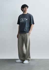 Jeune homme aux cheveux bouclés portant un t-shirt graphique noir délavé, un pantalon beige ample et des baskets blanches, debout devant un fond clair uni.