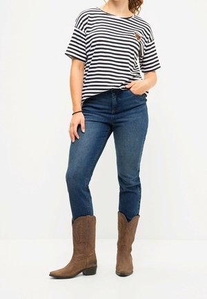 Kvinde iført sort- og hvidstribet t-shirt, blå jeans og brune cowboystøvler, stående med den ene hånd i lommen mod en hvid baggrund.